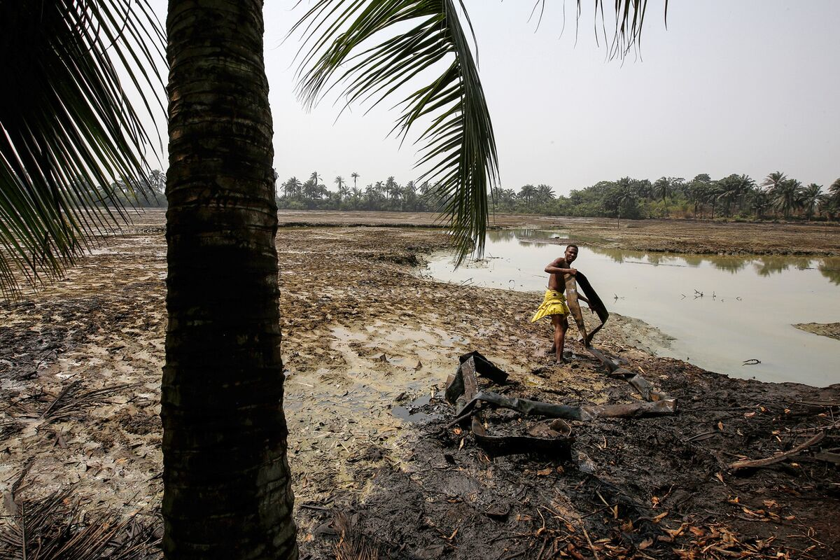 Man stands amid polluted water and debris in a rural area surrounded by palm trees, highlighting environmental impacts of resource extraction